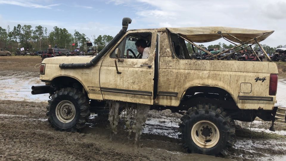 Mud-covered 1988 Ford Bronco with oversized tires and snorkel driving through mud.