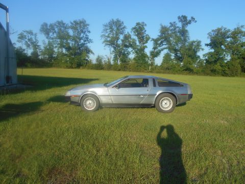 1992 Ford Bronco - A silver DeLorean DMC-12 is parked on a grassy field next to a building. The car is viewed from the side with the front wheel visible.