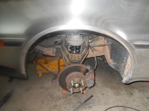 1992 Ford Bronco - The front suspension and brake assembly of a vehicle is visible, supported by a yellow hydraulic jack. The wheel hub and brake rotor are pr…