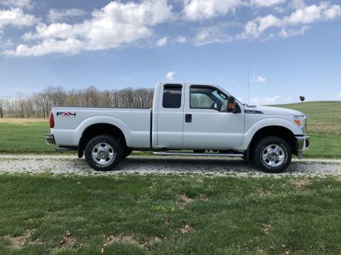 2011 Ford F-250 - A white Ford F-250 pickup truck is parked on a gravel road with a grassy field and trees in the background under a partly cloudy sky.