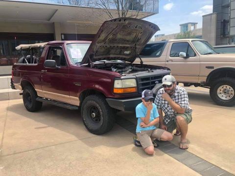 1996 Ford Bronco - A father and son examine the engine of a maroon Ford Bronco, with another truck in the background.