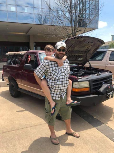 1996 Ford Bronco - A man carries a child on his back in front of a maroon SUV with its hood open.