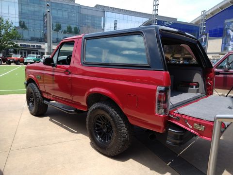 1996 Ford Bronco - A red 1996 Ford Bronco with a black top is parked outside a modern building, with its tailgate open.