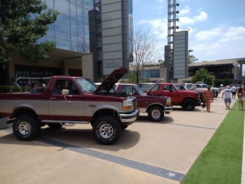 1996 Ford Bronco - A line of Ford Broncos are parked outside a modern building, with one prominently displayed in maroon and tan.