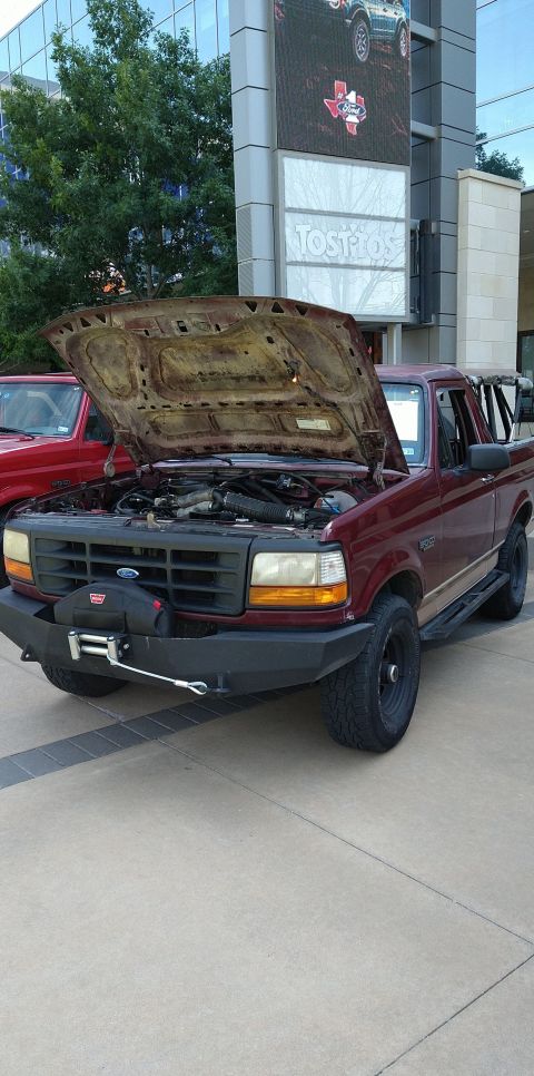 1996 Ford Bronco - A maroon Ford F-Series truck with its hood up is parked next to another red vehicle in front of a modern building.
