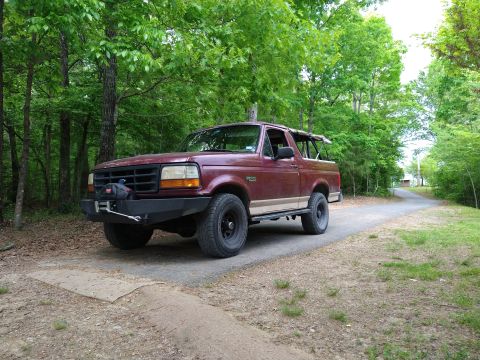 1996 Ford Bronco - A maroon 1996 Ford Bronco is parked on a road surrounded by trees.