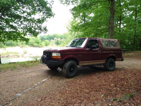 1996 Ford Bronco - A maroon Ford Bronco is parked on a dirt path near a body of water, surrounded by trees.