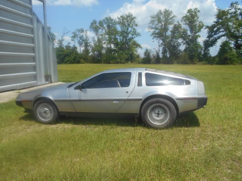 1992 Ford Bronco - A silver DeLorean DMC-12 is parked on a grassy field next to a corrugated metal structure. The car is viewed from the side, showcasing its…