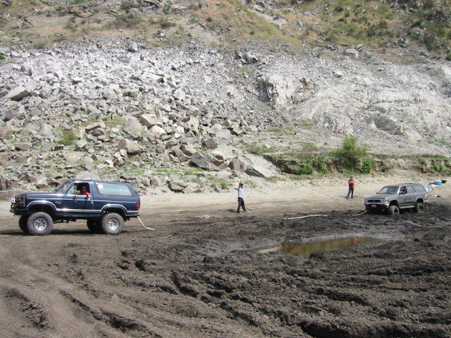 Blue lifted Ford Bronco with white top on muddy quarry terrain, gray SUV and people in background