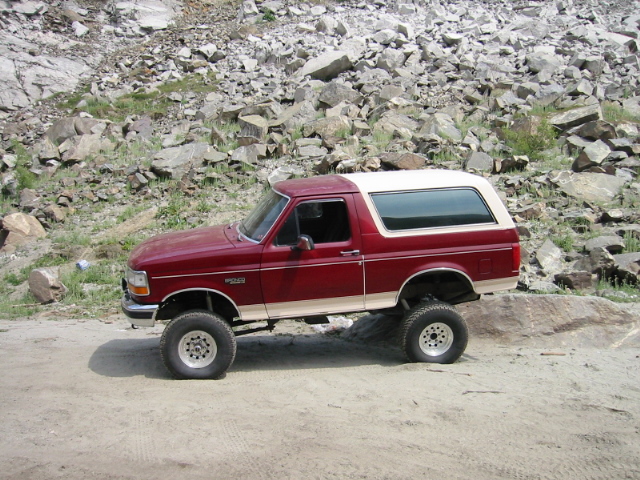 Maroon Ford Bronco with white top, lifted on off-road tires, parked by a rocky slope.
