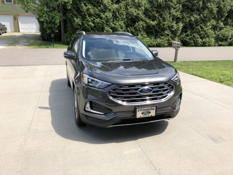 2019 Ford Everest - A dark gray Ford Edge SUV is parked on a concrete driveway in front of a tree line and a house.