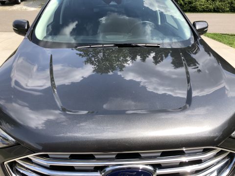 2019 Ford Everest - The front of a dark gray Ford Everest SUV is shown, reflecting the sky and trees on its hood. The vehicle's chrome grille and Ford emblem a…
