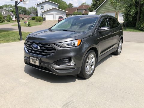 2019 Ford Everest - A dark gray Ford Edge is parked on a concrete driveway in front of a suburban house. The front of the SUV is angled towards the camera, sho…