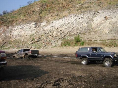 1985 Ford Bronco - A Ford Bronco tows a stuck pickup truck from a muddy area with a rocky hillside in the background.