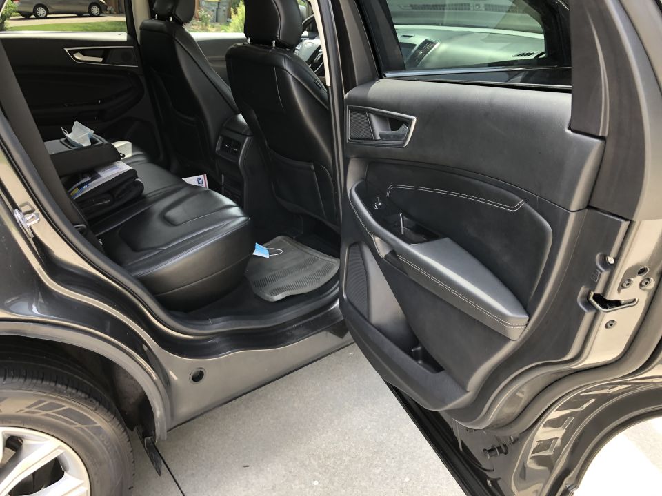 Interior of a dark SUV, rear seats and driver's door panel visible. Face mask on floor mat.