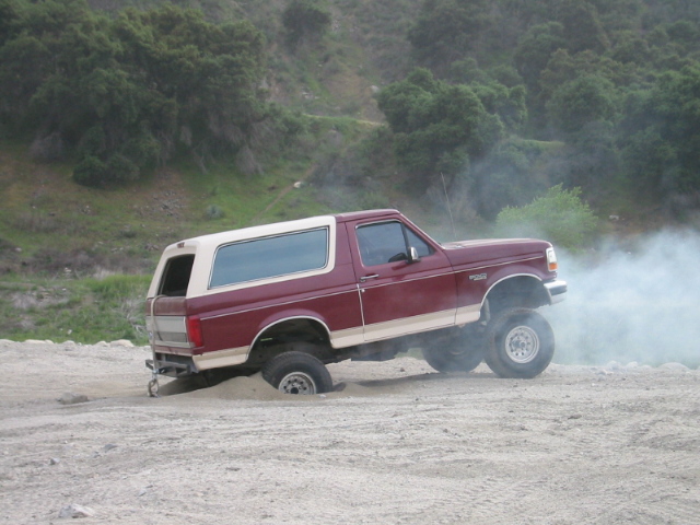 Maroon lifted Ford Bronco with white top spinning tires in dirt, side view