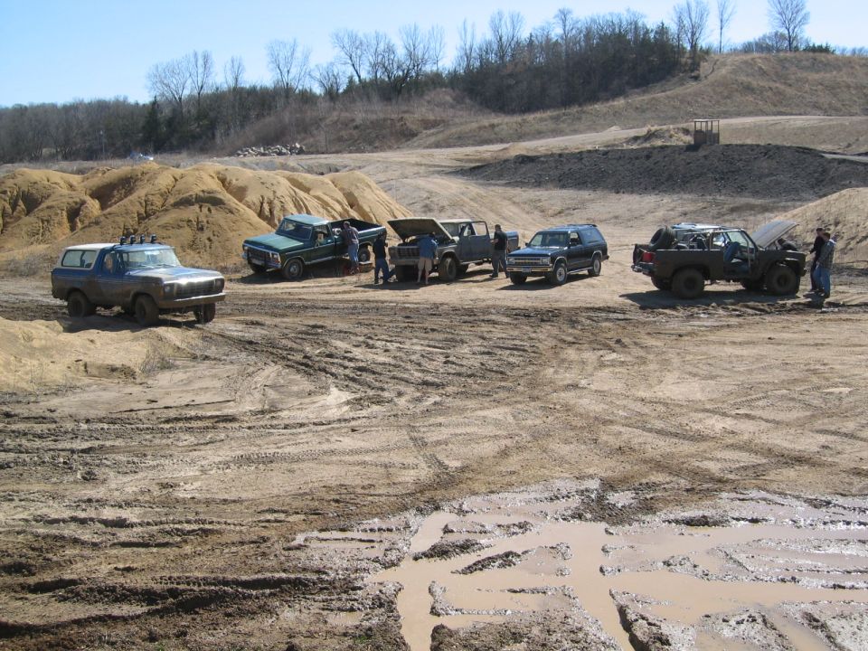 Group of trucks and SUVs parked in a muddy off-road pit, people gathered, hills and bare trees behind