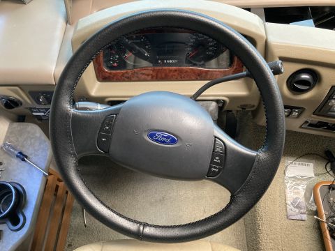 2015 Ford F-550 - Ford steering wheel with blue oval logo and steering-wheel cruise/audio control buttons, instrument cluster and tan dashboard behind it.