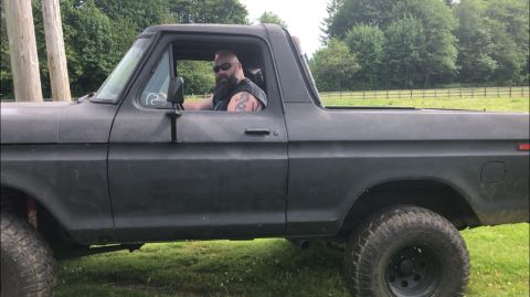 1978 Ford Bronco - A man sits behind the wheel of a dark-colored Ford Bronco, parked on a grassy field.