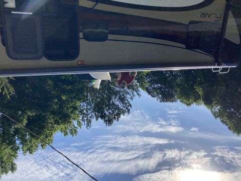 2015 Ford F-550 - A person stands next to a large RV with the name "FLAIR" visible on the side. The RV is parked outdoors with trees and a bright blue sky wi…
