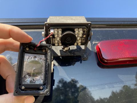2015 Ford F-550 - A person's hand holds the detached cover of a backup camera, revealing condensation inside. The camera itself, mounted on a dark vehicle, i…