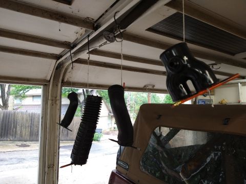 1996 Ford Bronco - Several black automotive intake parts are hanging from the ceiling of a garage, possibly being painted or cleaned. A portion of a tan soft-…