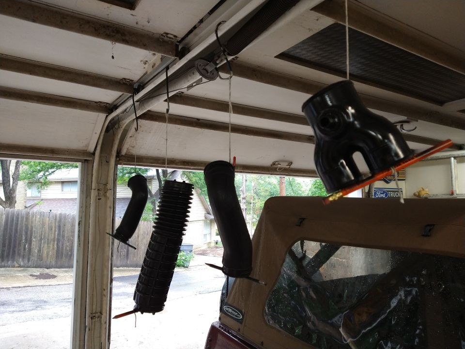 Black automotive intake parts hanging in a garage with a tan soft-top vehicle below.