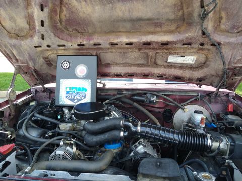 1996 Ford Bronco - The open hood of a maroon vehicle reveals a detailed engine bay with a sign for the "Bishop Dunne Car Show" prominently displayed.