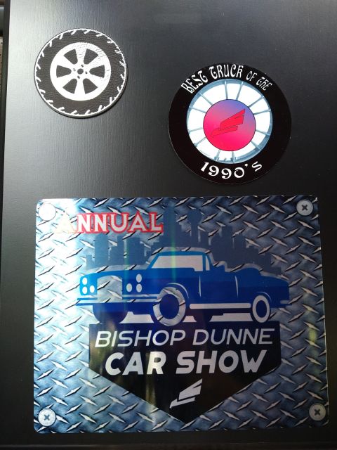 1996 Ford Bronco - Three stickers are affixed to a dark surface. The largest sticker is for the Bishop Dunne Car Show and features a blue truck. The other two…