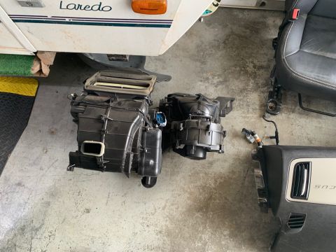 2008 Ford Focus - Two black automotive HVAC heater cores and blower motors are laid out on a concrete floor next to a black car seat and a section of a dashb…