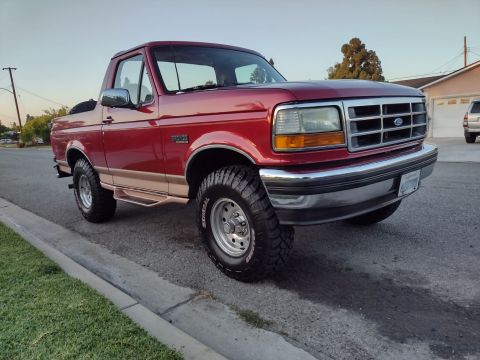 1994 Ford Bronco - This red Ford Bronco is parked on a residential street with a house and trees in the background.