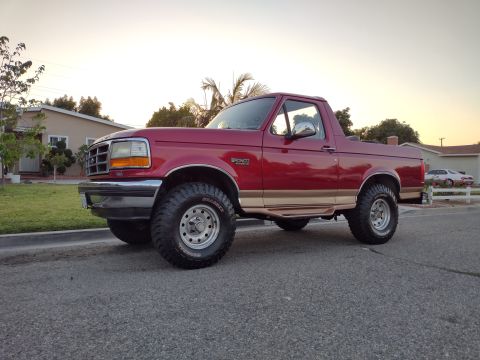 1994 Ford Bronco - A red Ford Bronco with tan trim and lifted suspension is parked on a street in front of a suburban house.