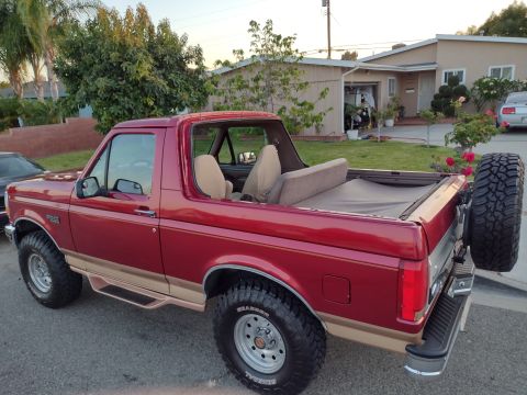 1994 Ford Bronco - Two-tone red over tan full-size Ford Bronco with the rear top removed, spare tire on tailgate, parked in a residential driveway.