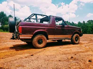 1996 Ford Bronco - This maroon Ford Bronco, equipped with a roll cage and spare tire, is parked on a dirt path with trees in the background.