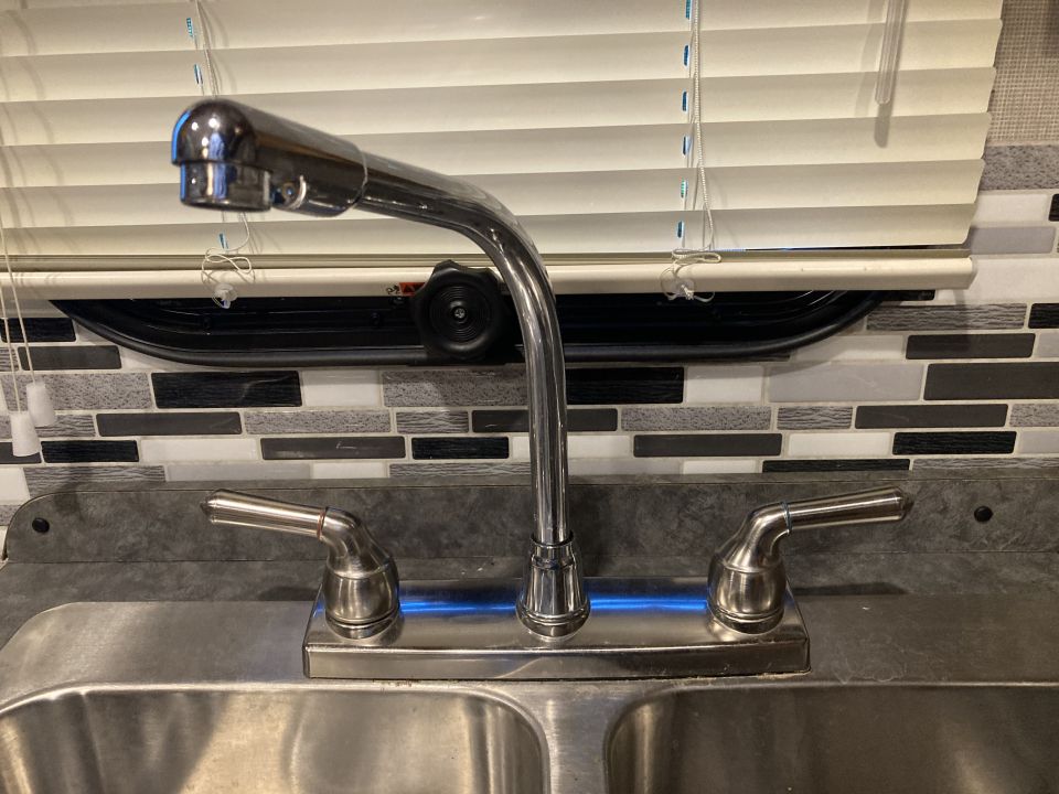 Kitchen sink faucet with two handles and a pull-out sprayer, mounted on a double sink. Blinds are visible in the background
