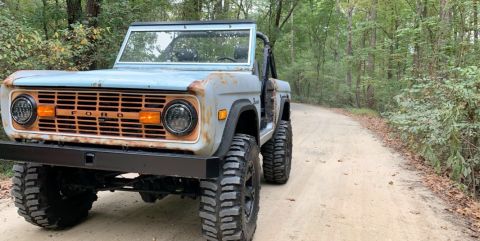 1976 Ford Bronco - This 1976 Ford Bronco features a distinctive rusty grille, large off-road tires, and a custom front bumper, seen on a wooded dirt road.