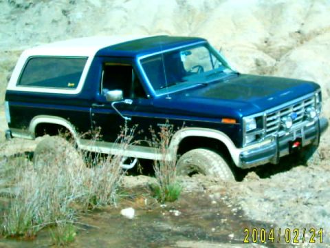 1985 Ford Bronco - A blue and white Ford Bronco navigates a muddy, off-road terrain, with its front wheels partially submerged.