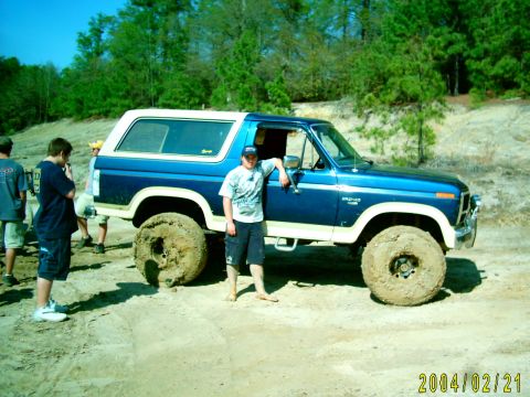 1985 Ford Bronco - A blue and white Ford Bronco sits in a dirt area with several people nearby.