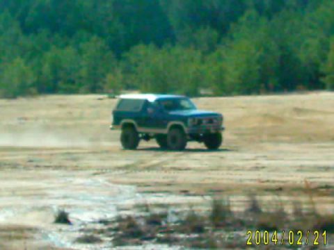 1985 Ford Bronco - A blue SUV drives across a dusty, open area with trees in the background.