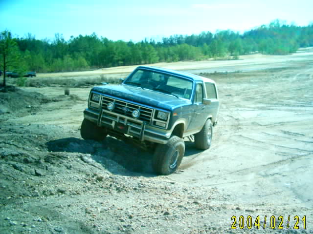 Blue 1985 Ford Bronco driving on a dirt hill with trees in the background