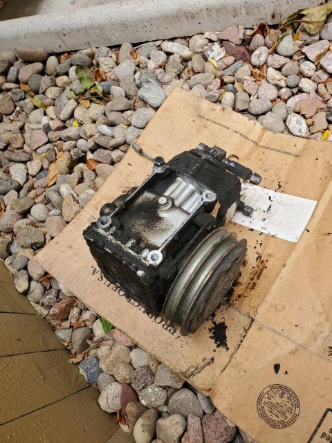 1996 Ford Bronco - This is a used automotive AC compressor, covered in grime and oil, resting on a piece of cardboard.