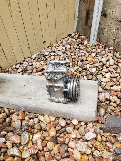 1996 Ford Bronco - An air conditioning compressor sits on a concrete ledge surrounded by rocks and a wooden fence.