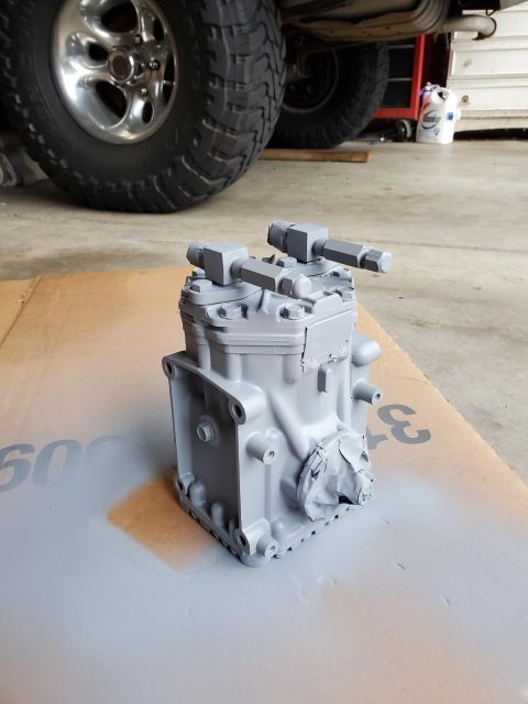 1996 Ford Bronco - A gray air conditioning compressor is shown on a piece of cardboard, with the tire and undercarriage of a vehicle visible in the background.