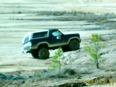 1985 Ford Bronco - A dark blue and white SUV with off-road tires navigates a dirt incline in a barren landscape.