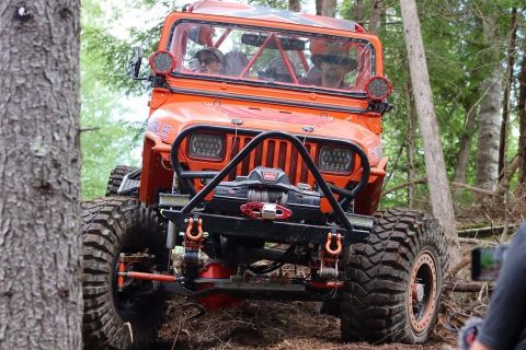 1992 Jeep Wrangler - Modified orange Jeep Wrangler navigates a muddy, wooded path with large off-road tires.