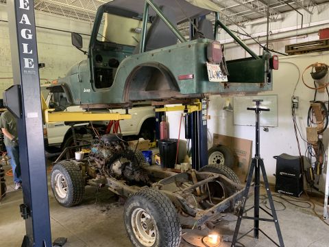 1979 Jeep CJ5 - A green Jeep CJ5 body is suspended on a lift, with its chassis and engine components laid out below in a workshop setting.