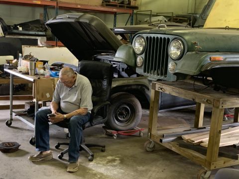 1979 Jeep CJ5 - A man sits in a chair next to a green Jeep CJ undergoing repairs in a workshop.