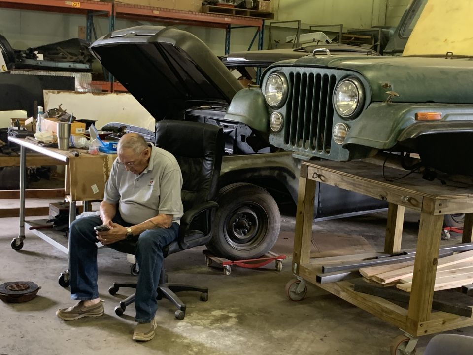Green Jeep CJ on a workbench, hood open, with a man sitting in a chair nearby