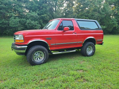 1995 Ford Bronco - This red Ford Bronco is shown from the side, parked on a grassy lawn with trees behind it.