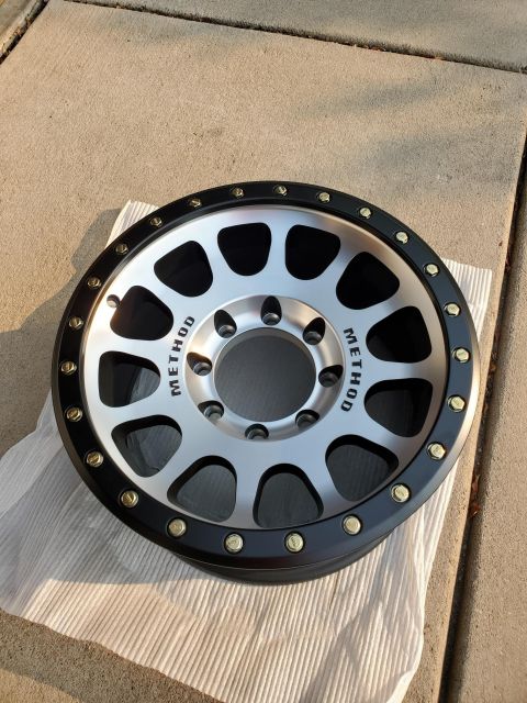 1996 Ford Bronco - A single Method brand wheel with a silver face and black outer lip is displayed on a white cloth on concrete.